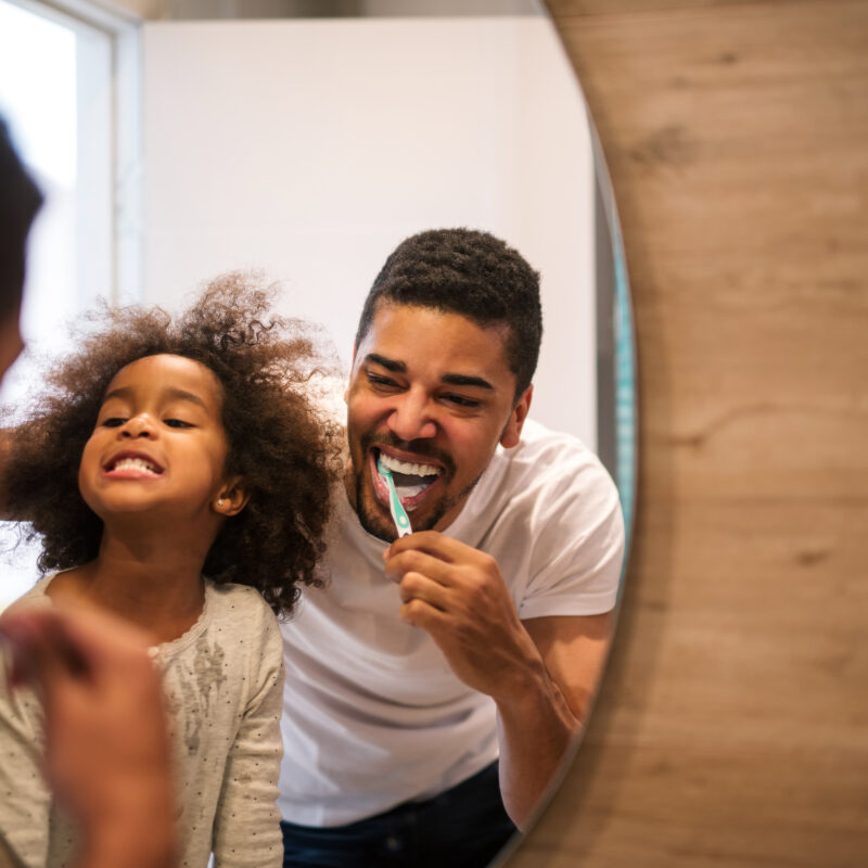 Father and daughter brushing teeth together in mirror.